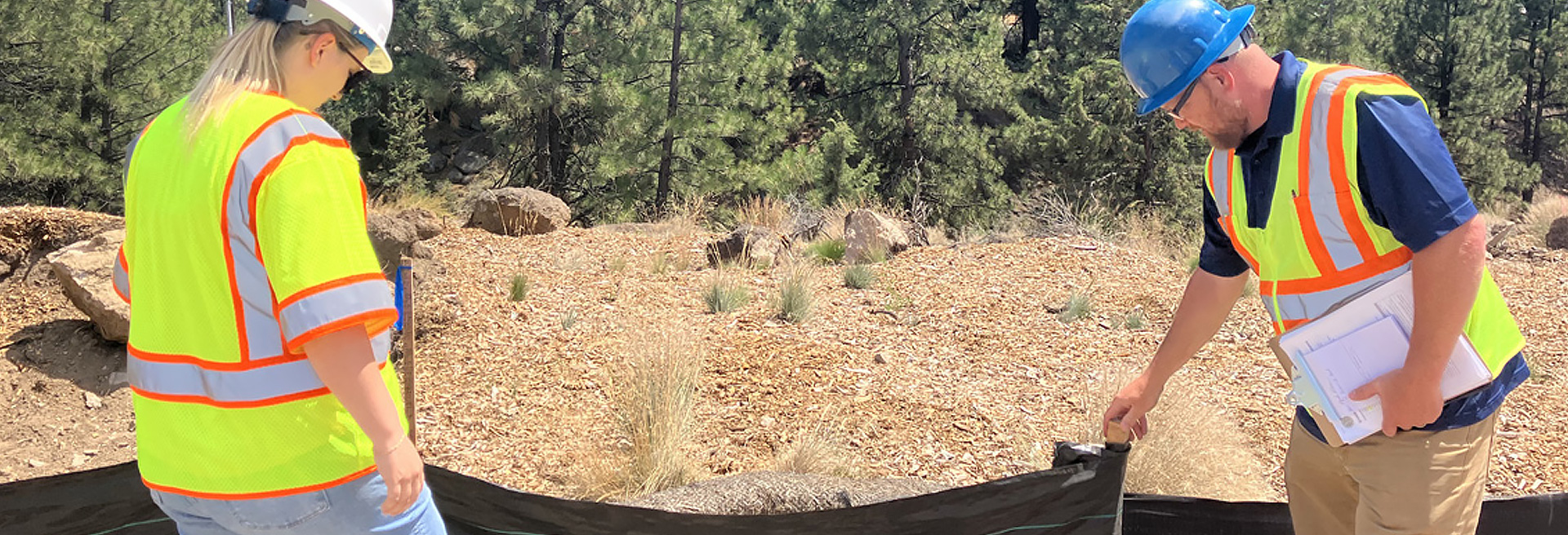 A man and a woman inspector wearing hardhats and safety vests inspection a temporary fence on a construction site