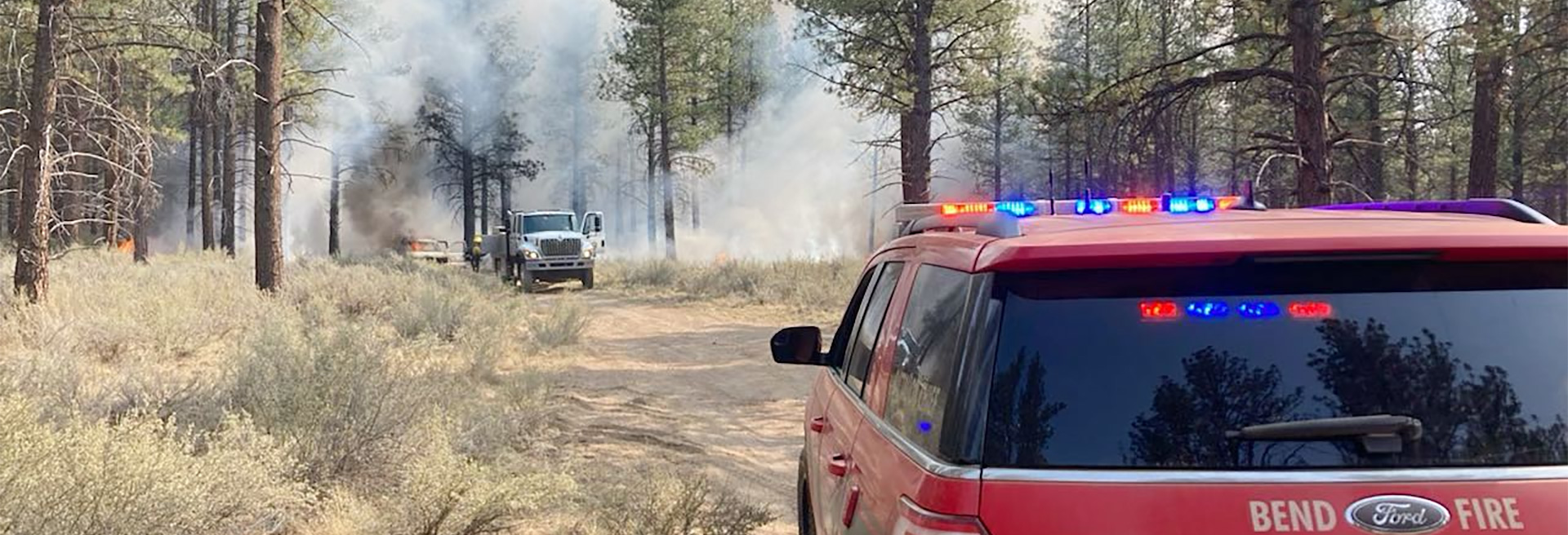 Red fire vehicle from the City of Bend on scene of a wildfire in a forest. Trees and brush all around, smoke and fire coming from the ground.