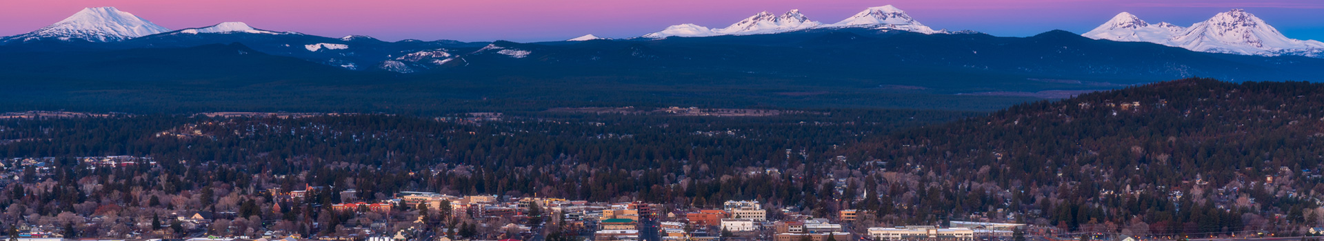 Overhead of Bend looking West towards the cascade mountains at late sunset. The sky is dark purple and the city is bursting with lights.