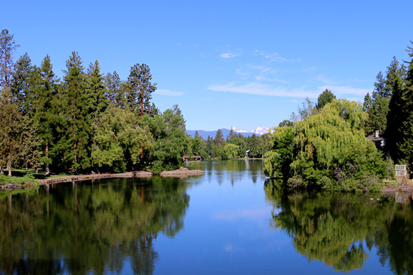 Mirror Pond with Drake Park to the left. Water is like glass reflecting the landscape and mountains on a blue sky day.