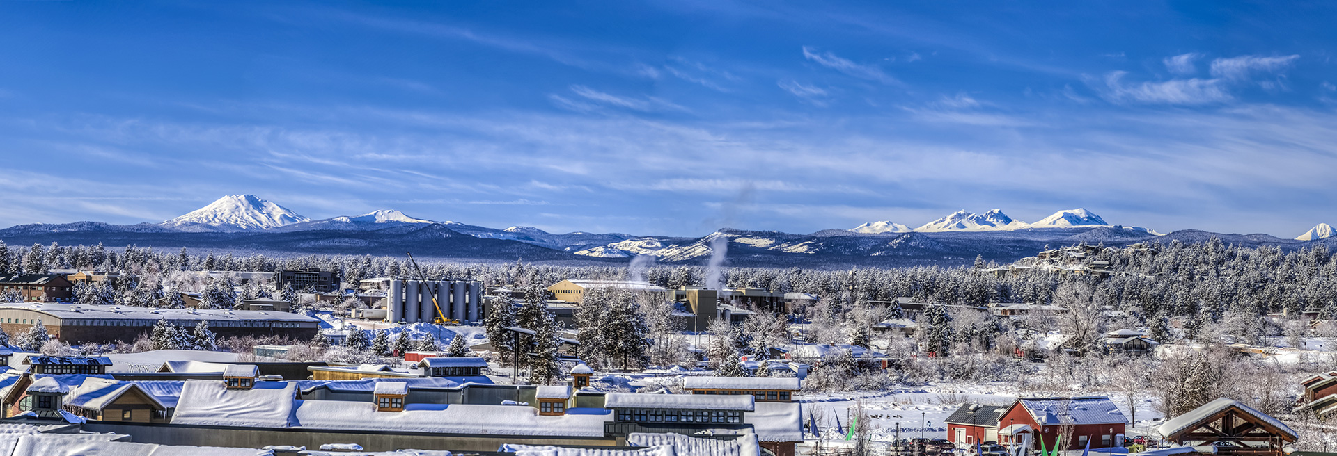 Overhead shot of Bend looking west in winter, sky is blue and the Cascade Mountains covered in snow. Businesses in the foreground.