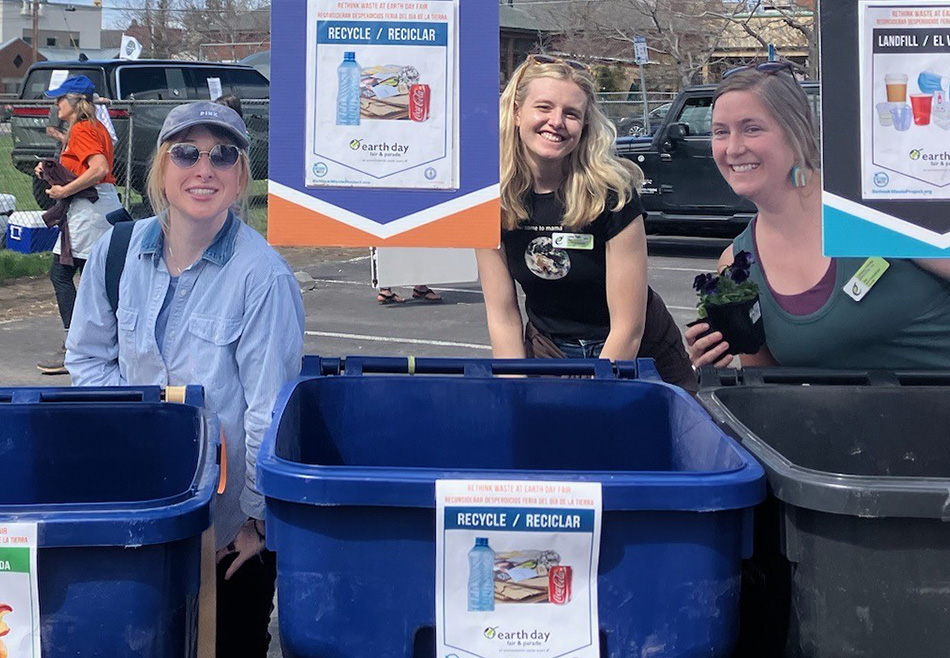 People behind recycling bins. Bins are blue and gray. Signs on each bins indicating what type of material belongs in each bin.