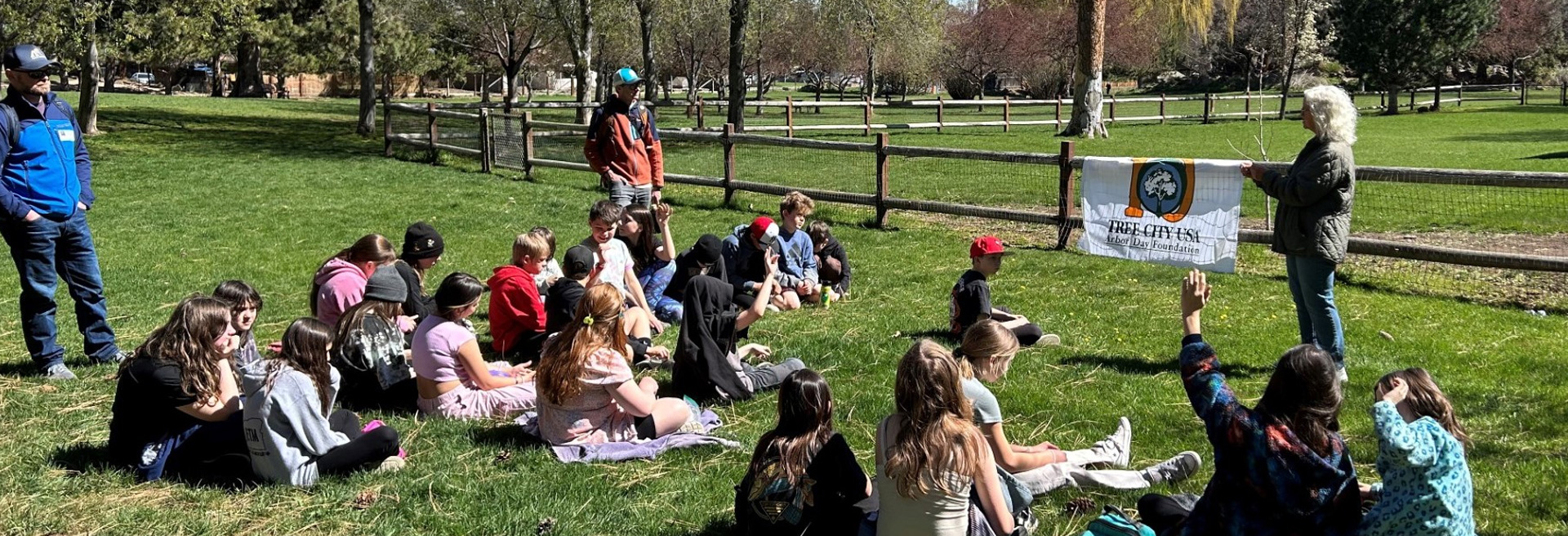 Teachers teaching children in a park. Teachers are standing, children are sitting. Grass is green and trees border the park.