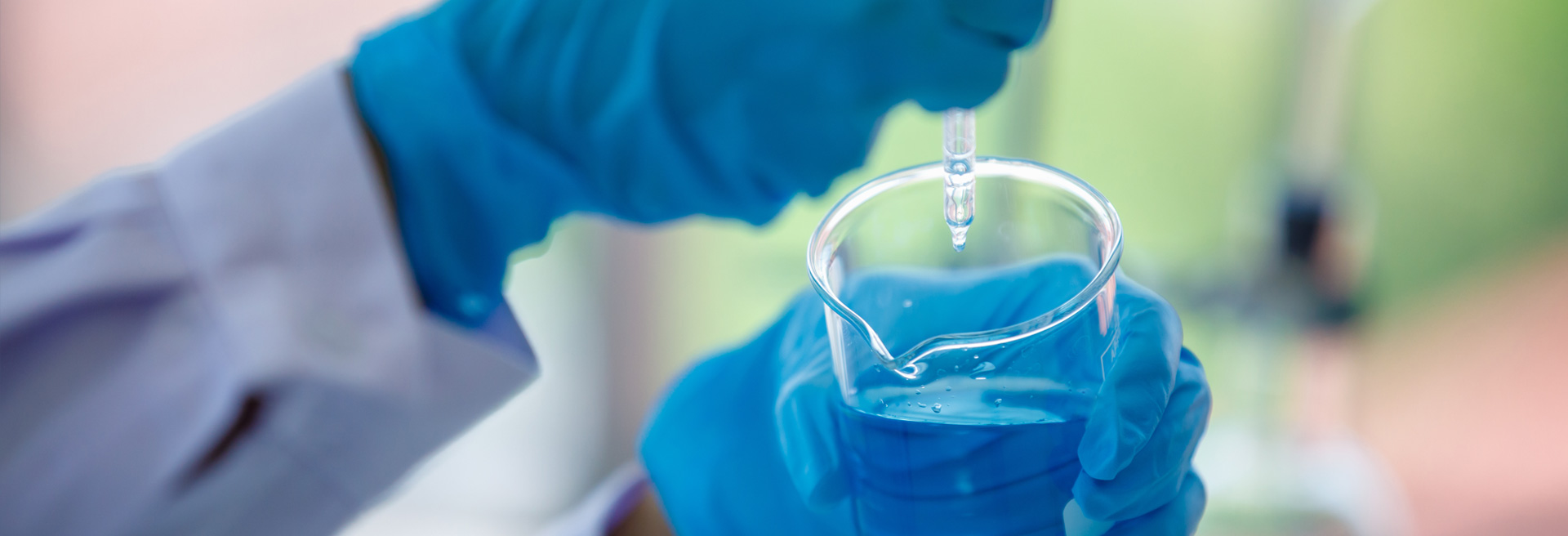 Person working in a laboratory with a white lab coat on, wearing blue gloves, using an eyedropper to drop water into a beaker. 