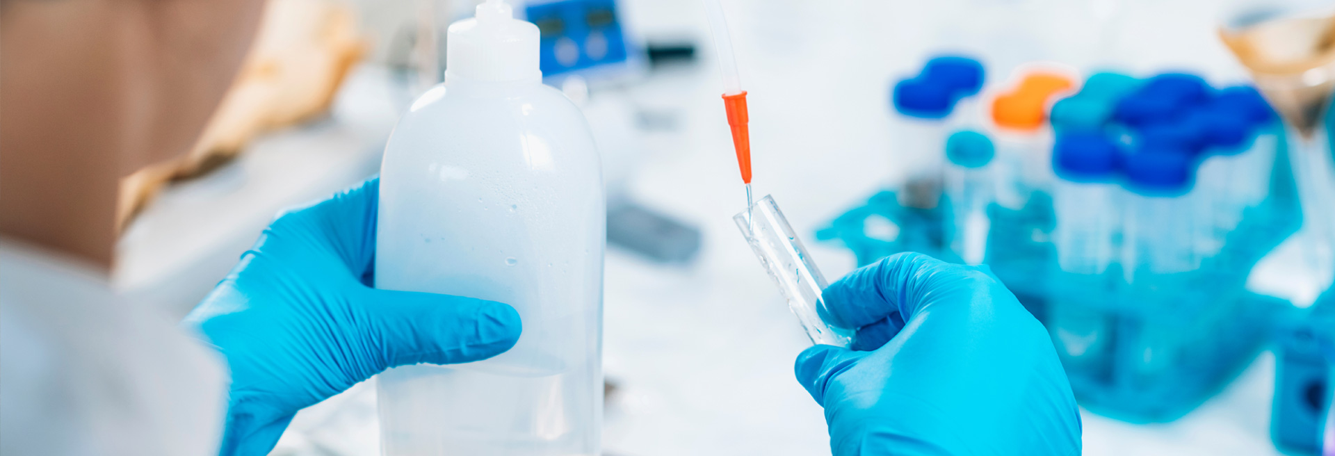Lab technician wearing blue gloves and a white coat, checking water from a bottle and pouring it into a test tube. 