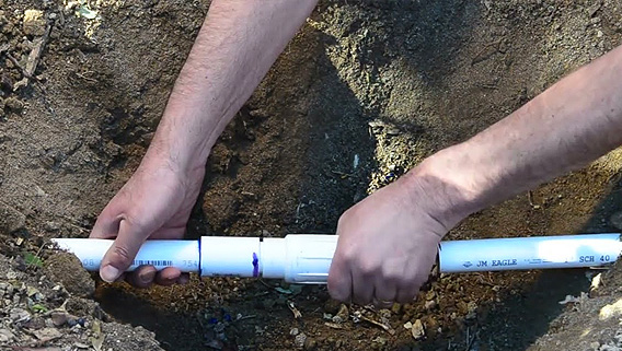 Hands repairing a broken white PVC pipe in a dirt hole.