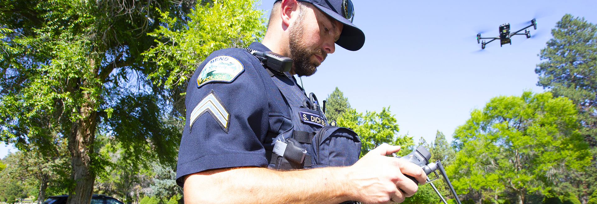 Police office holding a controller for a drone, the drone flies overhead in the background.