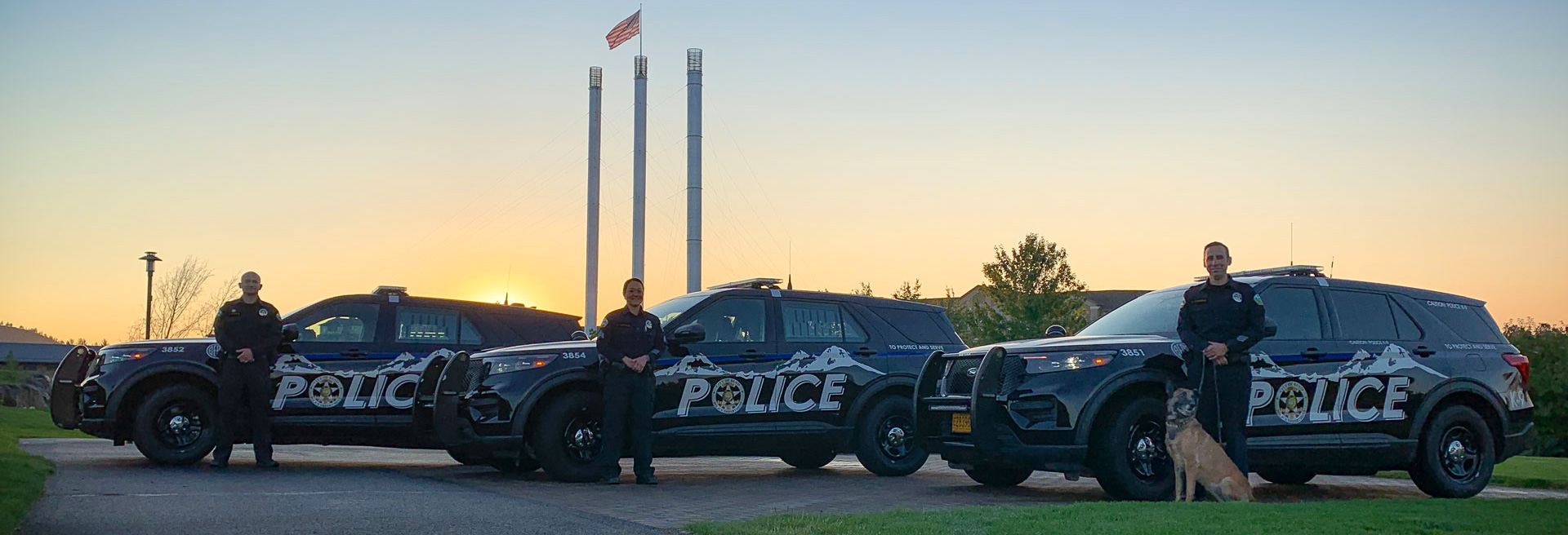 Three police vehicles parked with officers in front of them. The Old Mill District smokestacks at sunset are in the background.